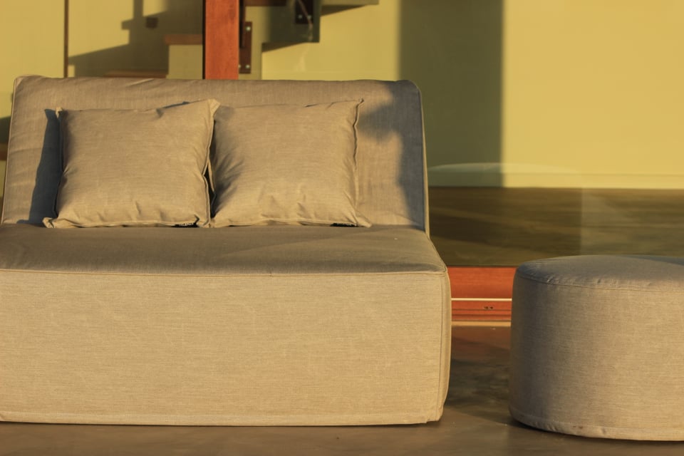 Sillón gris con almohadones y puff redondo en un patio con ventanal